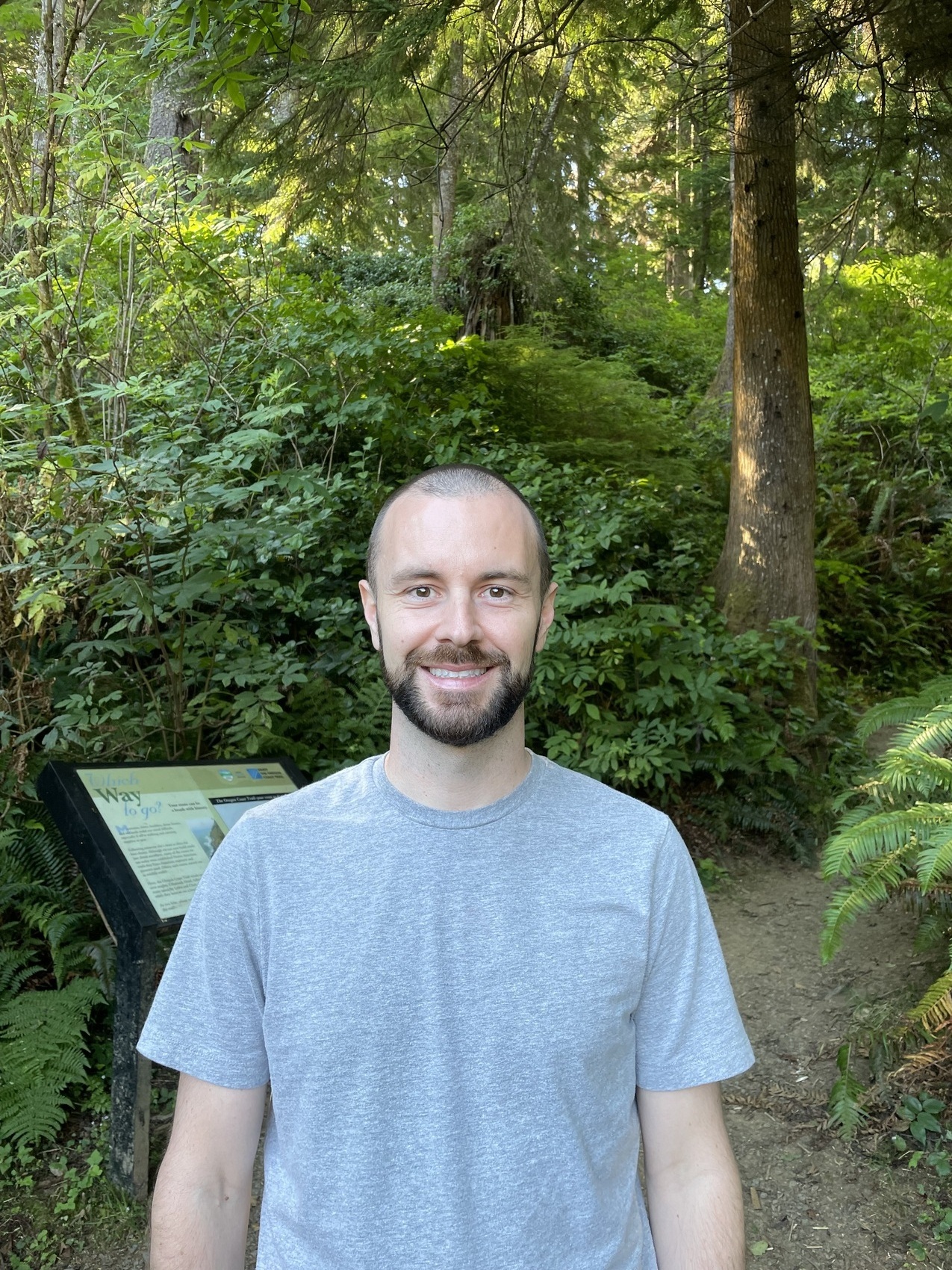 Professional photo of Andrew Buckley, a white bald man with a black beard and mustache. He is wearing a grey t-shirt and smiling, open-mouthed, in front of a backdrop of trees and shrubbery in a forested setting. 