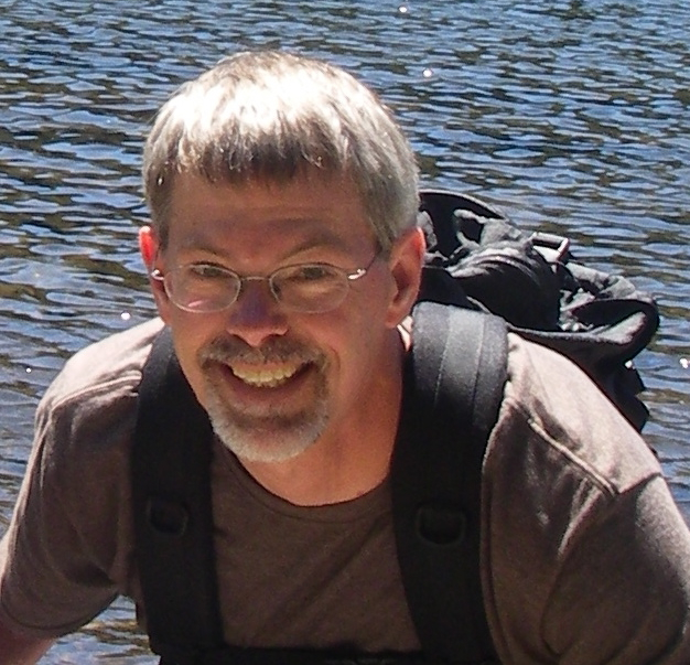 Headshot of Dirk Iwata-Reuyl, he is a white man with short grey hair and a beard and mustache. He is wearing a black backpack, a grey t-shirt, and clear framed glasses. He is smiling and leaning forward towards the camera with water as a backdrop.