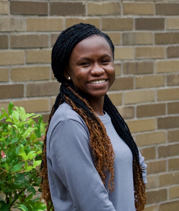 A headshot of Abigail Donkor, a black woman with long black hair in braids. She has blonde highlights. She is wearing a light grey-blue long sleeve shirt and smiling with teeth. She is facing the camera with her body posed towards the right, standing in front of a brick wall background with a small green bush to her left. 