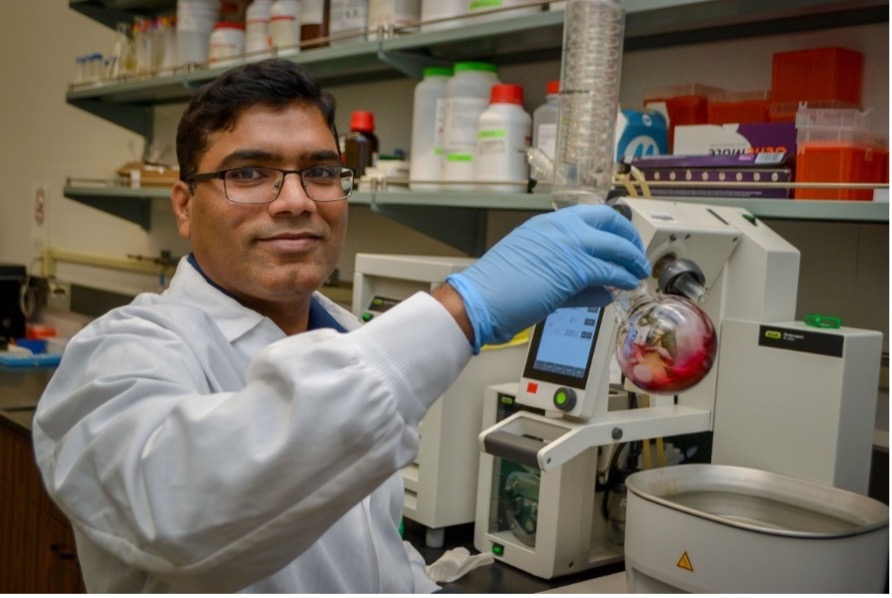 A professional portrait of Reddy Kancharla, a brown-skinned man with short black hair, wearing a white lab coat, blue gloves and black glasses. Kancharla is actively working in the lab space.
