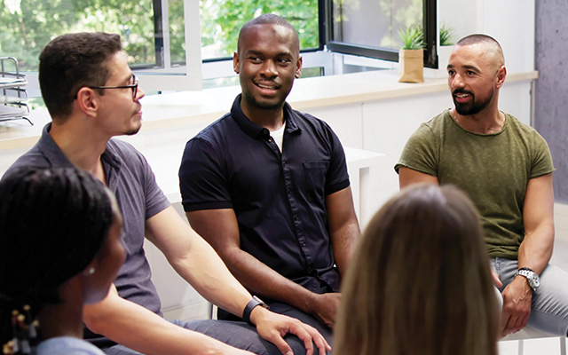 A group of diverse adults sit in a semicircle sharing their stories.