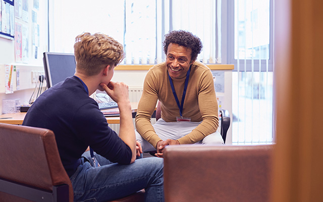 A school counselor with dark, curly hair and an ochre shirt smiles while advising a student with a blond undercut and dark blue clothes.