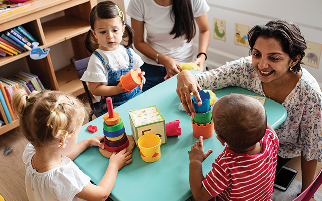 A teacher with short, dark hair smiles in a preK classroom as she helps children with blocks.