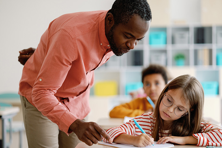 A Black male teacher in a salmon button-down and khaki slacks assists a student with her classwork.