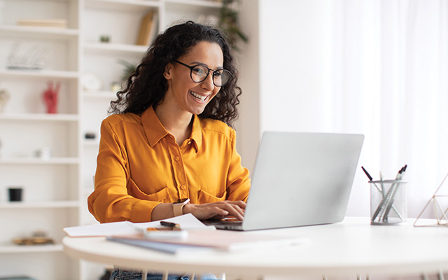 A graduate student with long, wavy black hair, black glasses and an orange shirt works on a laptop.