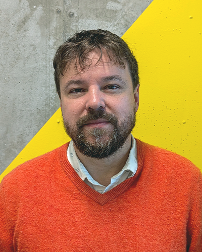 A headshot of Jesse Chapman, a subtly smiling Causasian man with short, dark brown hair and beard. He wears an orange sweater over a white button-down.