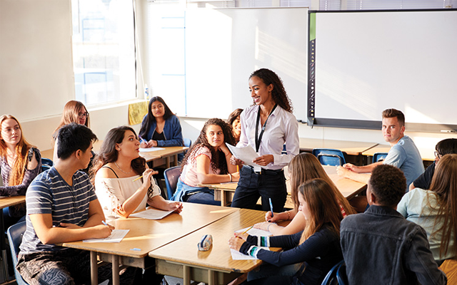 A teacher stands in the middle of a secondary school classroom talking with her students.