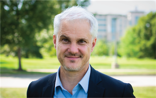 Photo of Jacob Wagner standing in front of some greenery and a distant building.