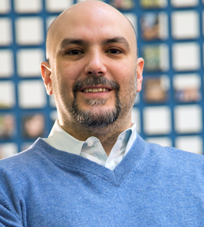 Photo of Luciano Berardi wearing a blue sweater and standing in front of a blue grate. 