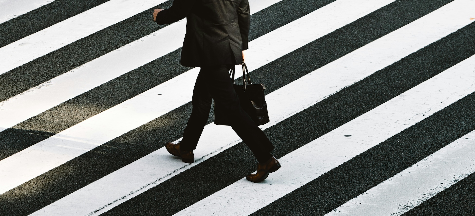 Photo by Ryoji Iwata, from Unsplash.com. Image of a man in a suit walking in crosswalk with briefcase.