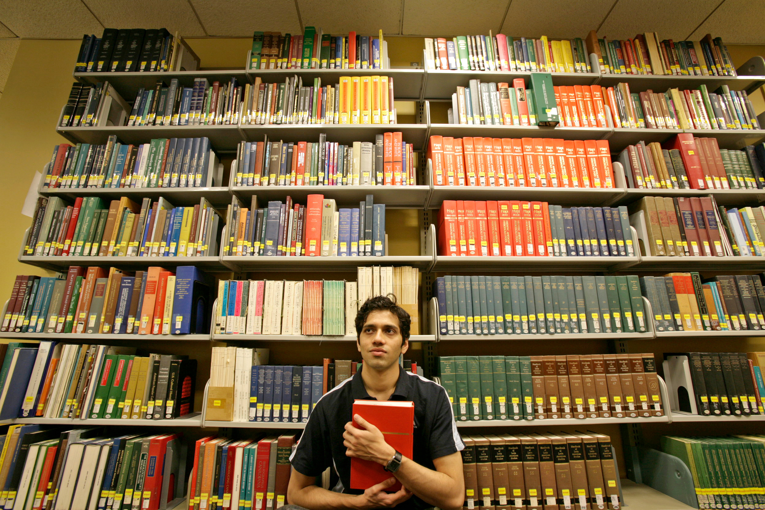 A student standing in front of tall library shelves holds a red bound book to their torso